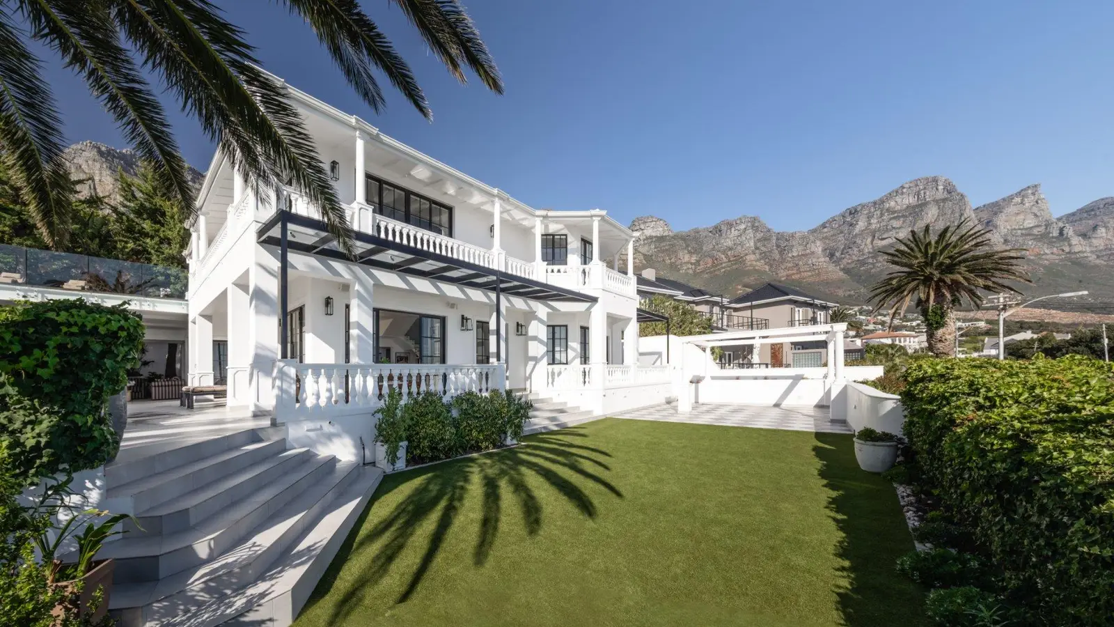 White two-storey house with balustraded balconies, a lawn and palms in front, mountains behind